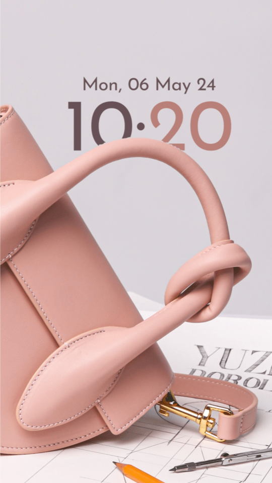 A pink leather handbag on a pale grey backdrop, its handle looping in front of a 10:20 clock in two shades of rose.